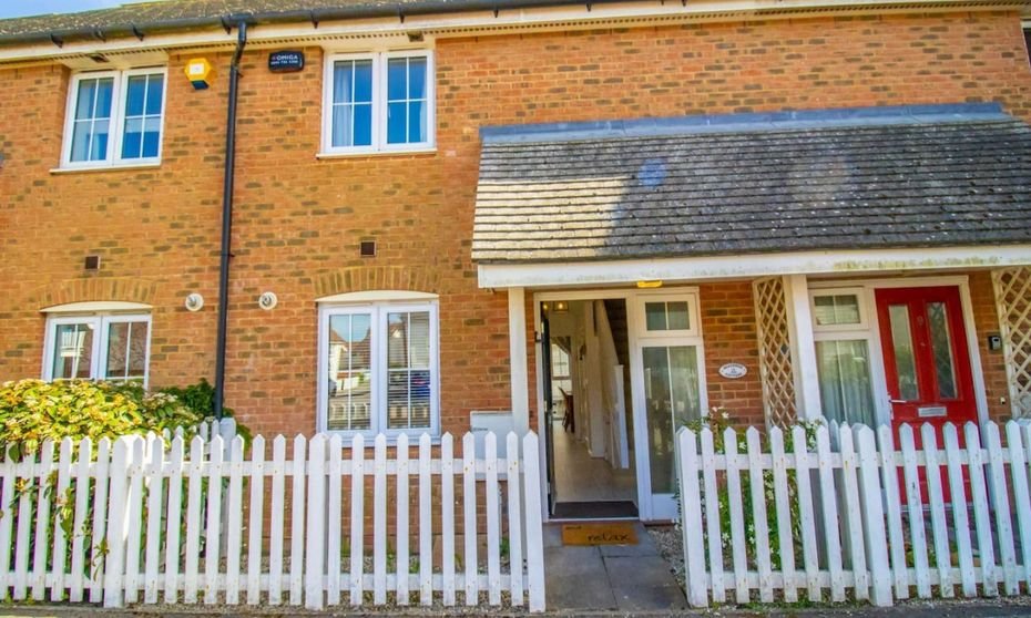 Beachside Cottage, a brick terraced holiday cottage with a white picket fence and red front door in Camber, East Sussex