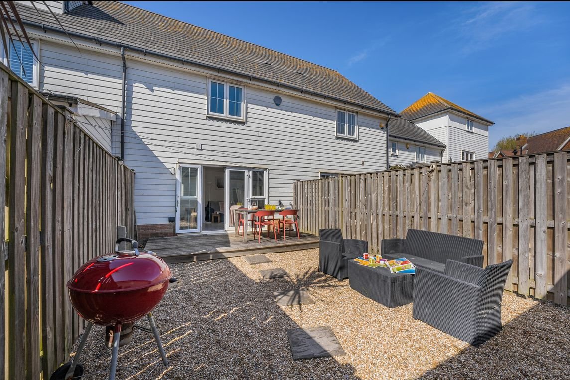 The private rear garden at Beachside Cottage with decking, French windows, outdoor dining table, rattan sofas and a BBQ on the pebbled area